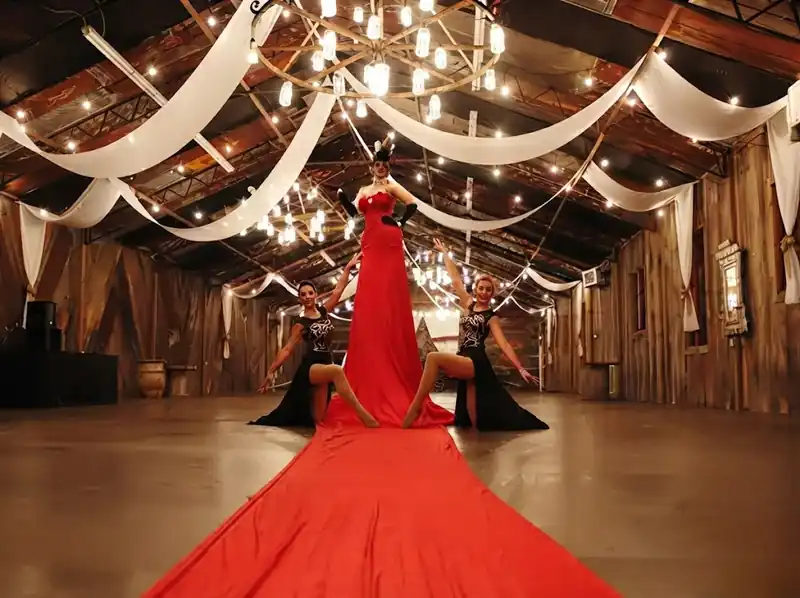 Mujer con vestido rojo de cola extra larga posando arrodillada, junto a dos bailarinas vestidas de negro, en un salón rústico decorado con luces colgantes y telas blancas.