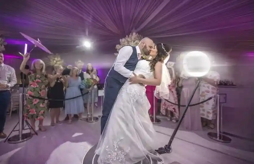 Novios utilizando un fotomatón 360 durante una boda, besándose sobre una plataforma giratoria mientras los invitados observan alrededor, con iluminación LED y ambiente festivo en interiores.