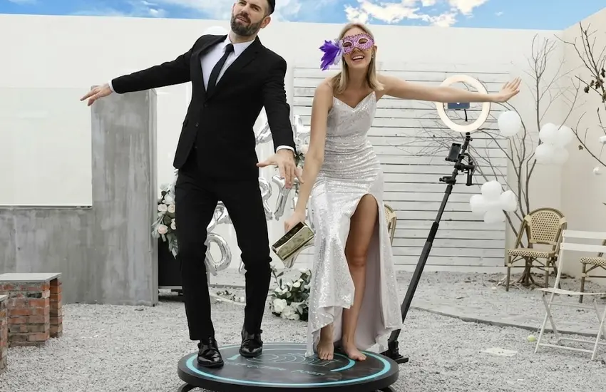 Pareja elegantemente vestida utilizando un fotomatón 360 en un evento al aire libre, posando sobre una plataforma giratoria con iluminación y cámara profesional para crear vídeos inmersivos.