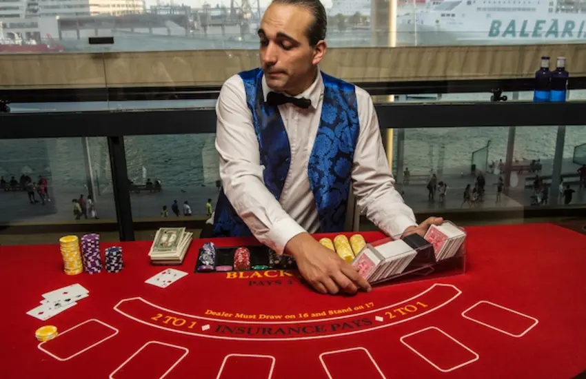 Crupier de casino vestido con chaleco azul repartiendo cartas en una mesa de blackjack durante un evento temático. Sobre la mesa se observan fichas de colores, cartas de juego y el tapete profesional de blackjack, con vistas al puerto y embarcaciones al fondo.
