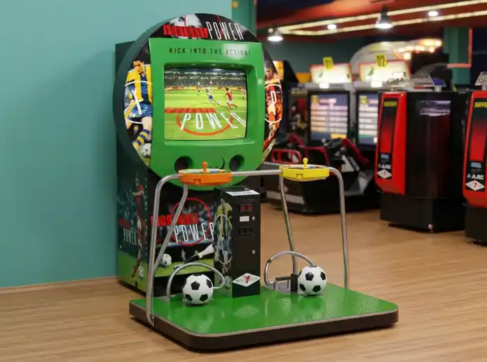 Máquina recreativa de fútbol interactiva con pantalla integrada, plataforma de juego y dos estaciones de control. Los jugadores utilizan balones y controles para participar en desafíos de fútbol virtual mostrados en la pantalla, combinando habilidad física y entretenimiento digital en una experiencia competitiva y divertida.  Pie de foto: