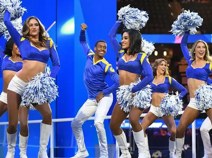 Texto alternativo: Un grupo mixto de animadores de los LA Rams (un hombre y cuatro mujeres) posa dinámicamente sobre un escenario azul brillante. Visten uniformes de mangas largas azul real con detalles amarillos y pantalones o pantalones cortos blancos con botas altas blancas. Las mujeres sostienen grandes pompones blancos, y el hombre en el centro levanta un puño. Todos sonríen y miran hacia adelante en una formación de baile. El fondo es un estadio de baloncesto interior con gradas borrosas.