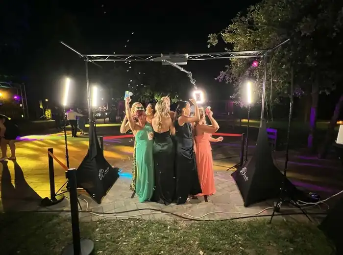 "Fotografía nocturna nítida y de alta resolución de un fotomatón 360 SKY en una fiesta al aire libre. Un grupo de cuatro personas de pie en una plataforma circular de metal con el logotipo 'SKY' posa y sonríe mientras un brazo de cámara automatizado con un anillo de luz gira alrededor de ellos. La estructura superior soporta el carril de la cámara. El fondo está decorado con luces de guirnalda y un área de DJ, bajo un cielo nocturno estrellado. La imagen es perfectamente clara y enfocada.