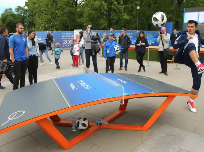 Participantes disfrutando de una partida de Teqball en un evento al aire libre, utilizando una mesa curva especial para realizar pases y remates con un balón de fútbol mientras el público observa la actividad.