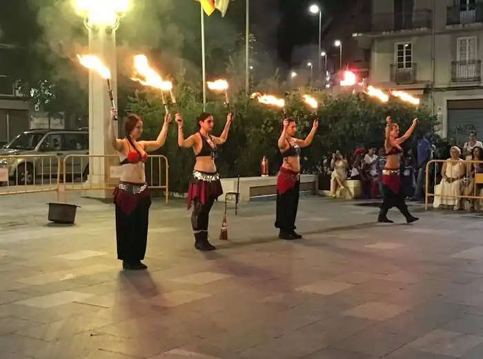 Grupo de cuatro artistas realizando un espectáculo de fuego al aire libre durante la noche, sosteniendo antorchas encendidas mientras el público observa desde el fondo de una plaza urbana.