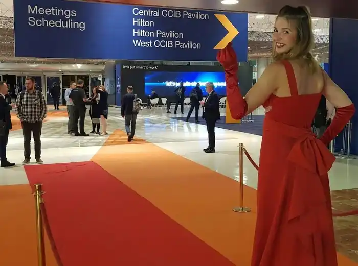 Mujer con vestido rojo largo y guantes a juego posando sobre alfombra roja en un centro de convenciones, con carteles de señalización azul al fondo.